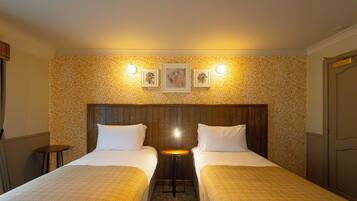 A hotel room with two beds, a small table, and a wall with patterned wallpaper and framed pictures.