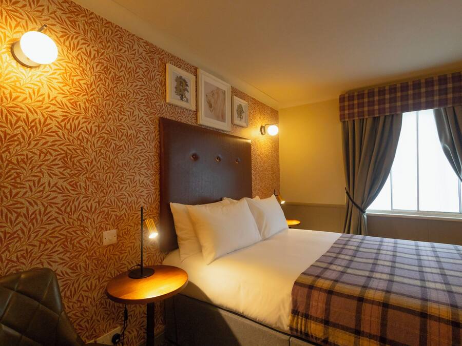 A hotel room with a patterned wallpaper, a headboard with lights, a bed with a plaid blanket, and a window with curtains.
