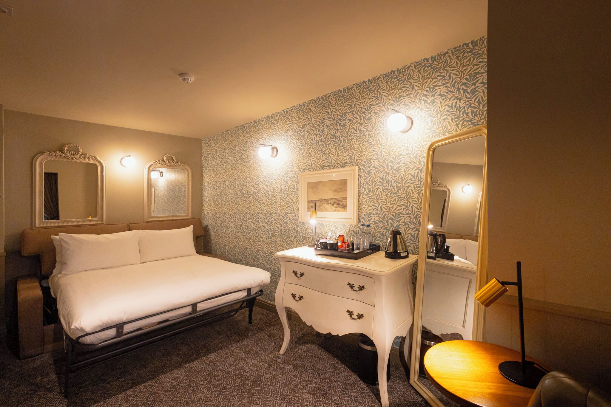 A hotel room with a bed, a bedside table with a lamp, a mirror, and a patterned wall.