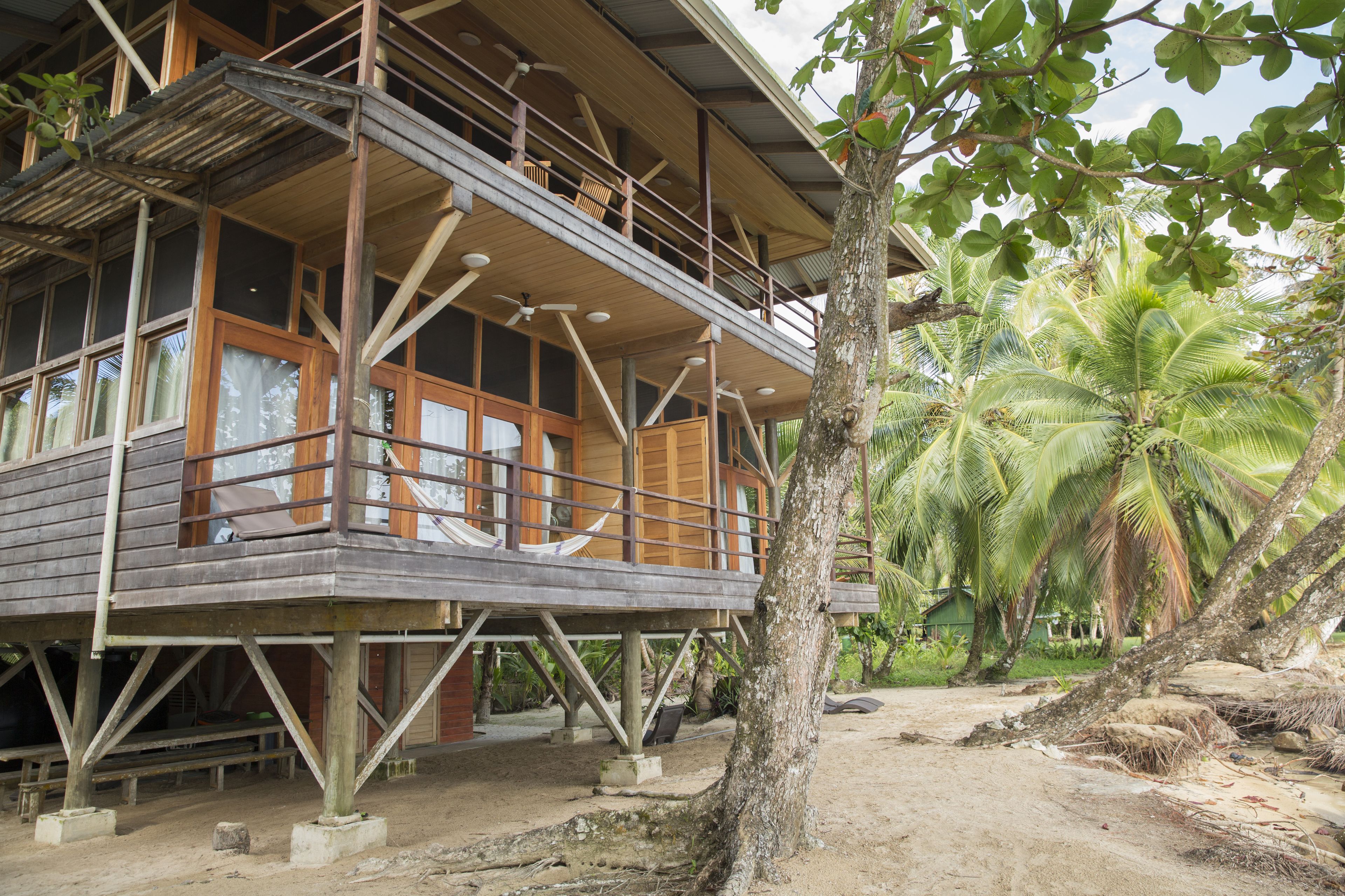 Una casa de madera con balcón, rodeada de palmeras y suelo arenoso.