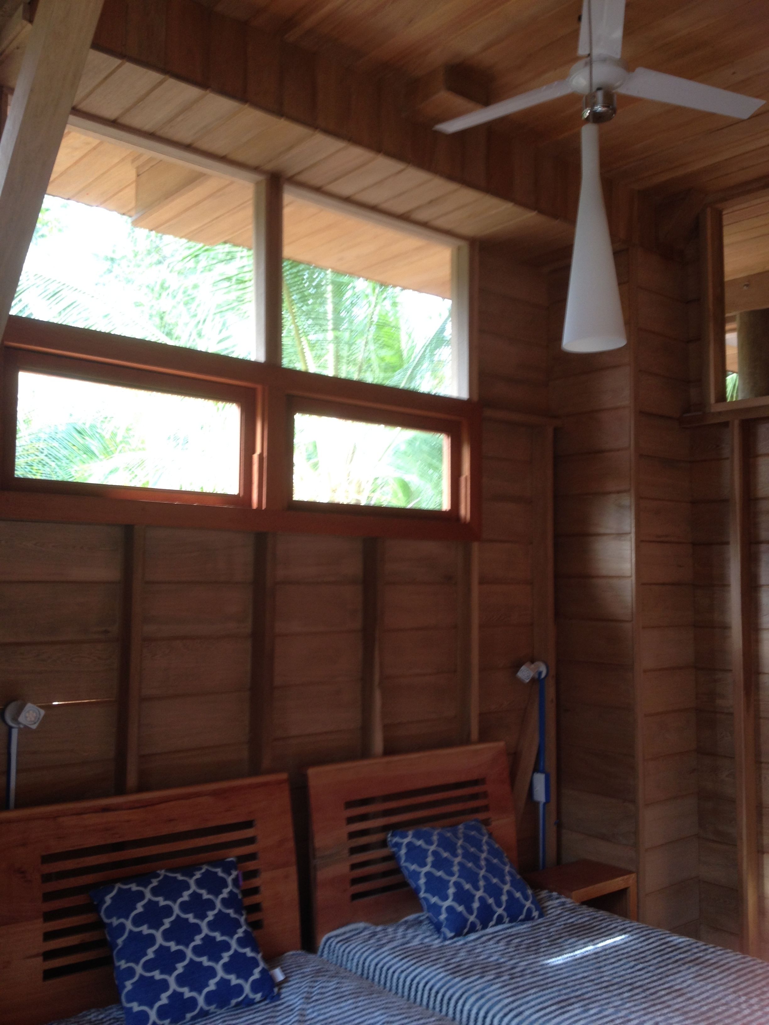 A wooden cabin room with a bed, a ceiling fan, and a window with a view of greenery.