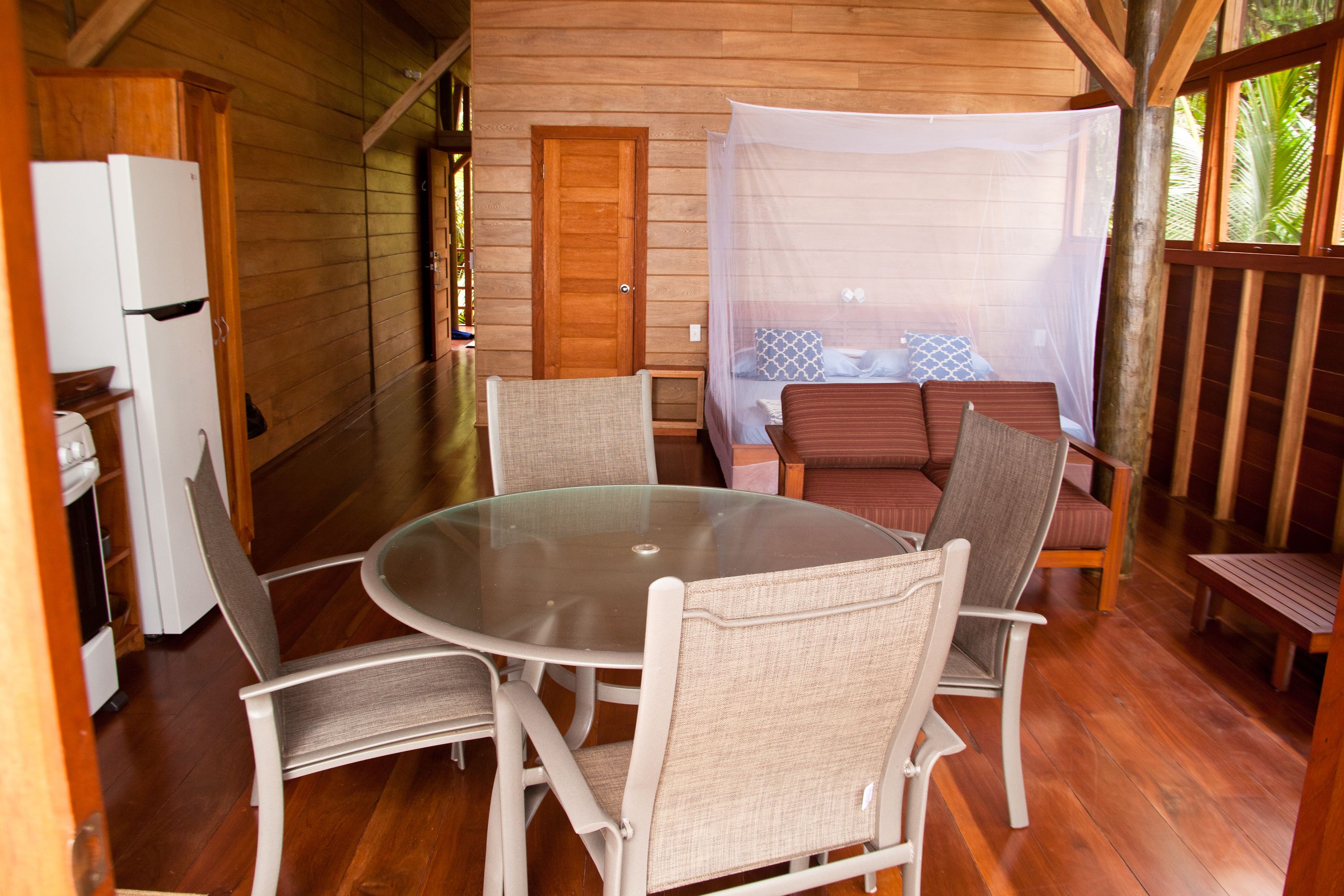 Una cabaña acogedora con una mesa redonda de cristal rodeada de sillas con cojines, una cama con mosquitero y una puerta de madera.