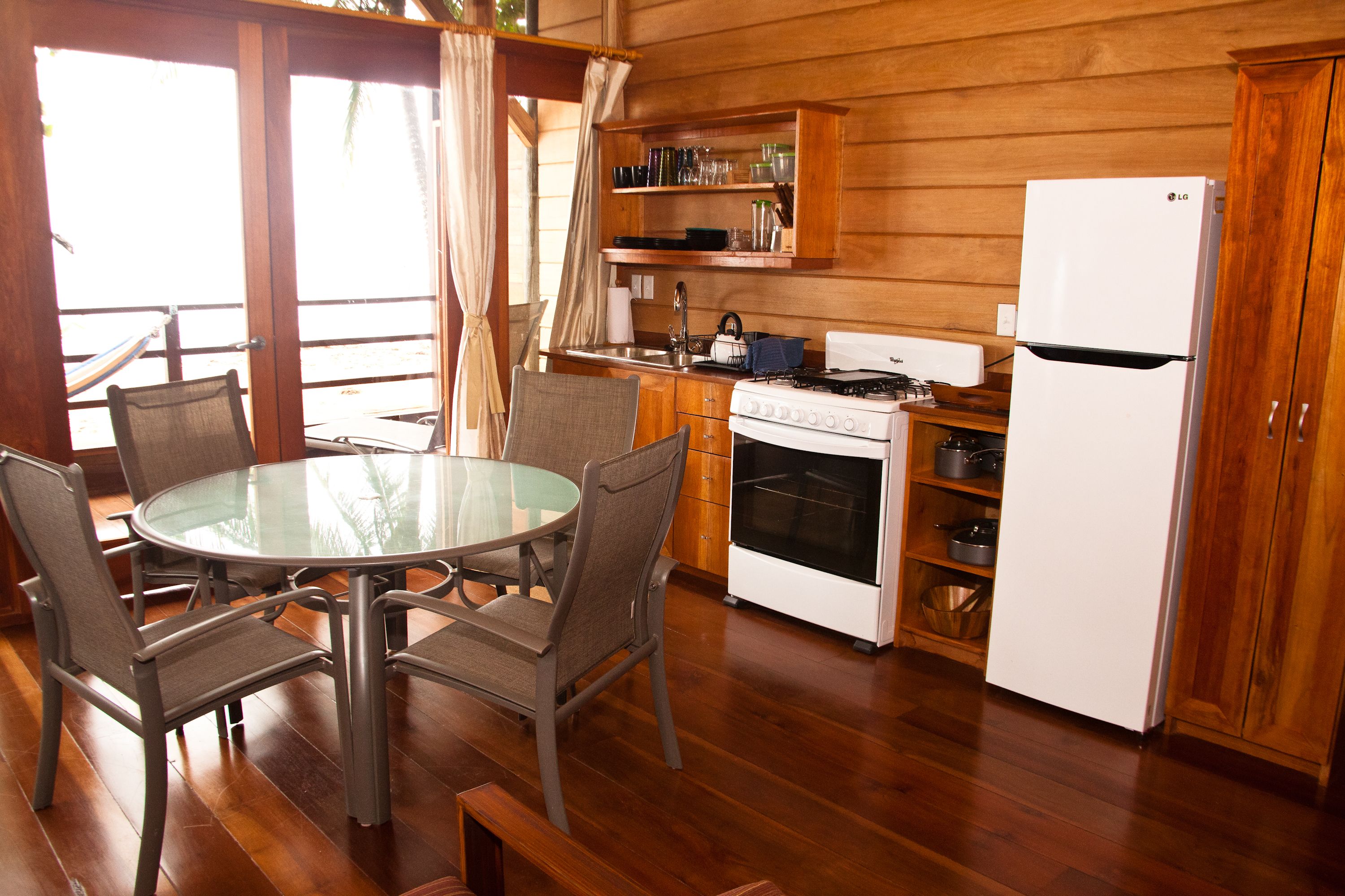 Een kleine keuken met een ronde glazen tafel en stoelen, houten keukenkastjes, een witte koelkast en een fornuis.