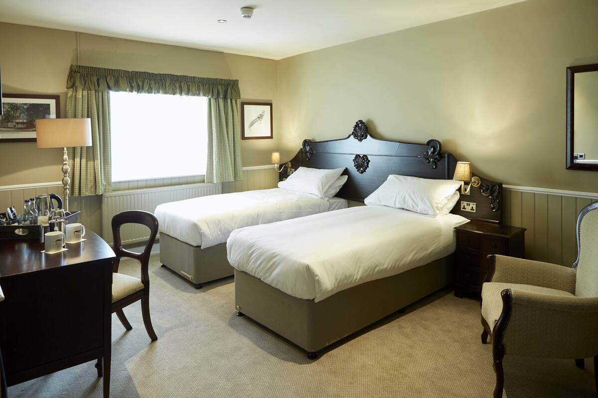 A hotel room with two single beds, a desk with a chair, a lamp, and a small table with cups.