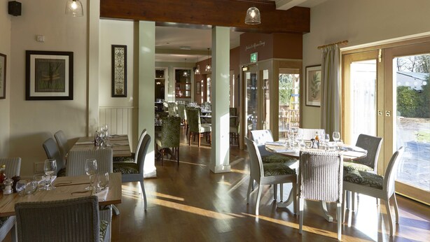 A restaurant with wooden tables and chairs, framed artwork on the walls, and large windows allowing natural light. - The Golden Pheasant (Knutsford)