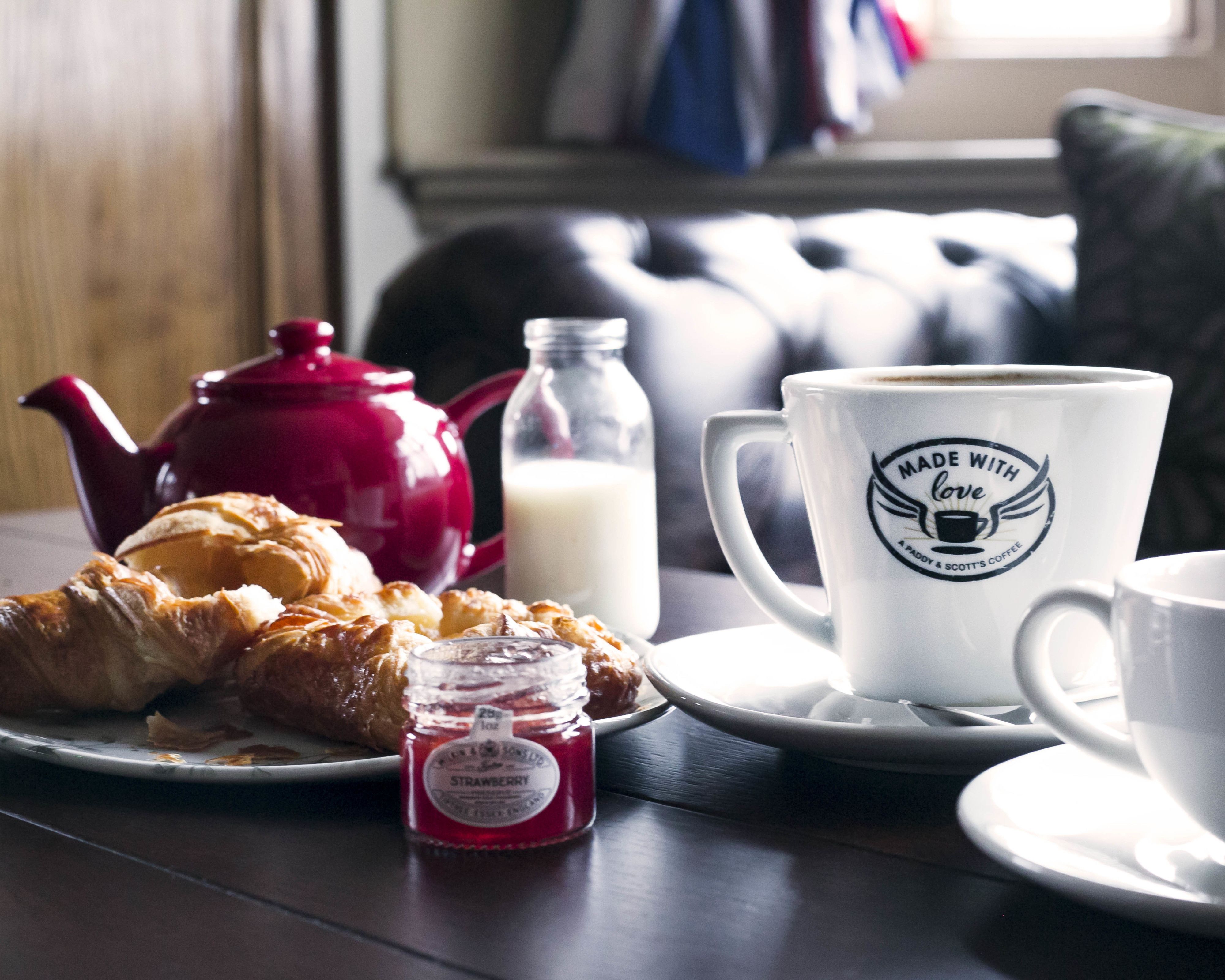 Una escena de desayuno con croissants, una tetera roja, una botella de leche, un tarro de mermelada de fresa y una taza de café con un logotipo.