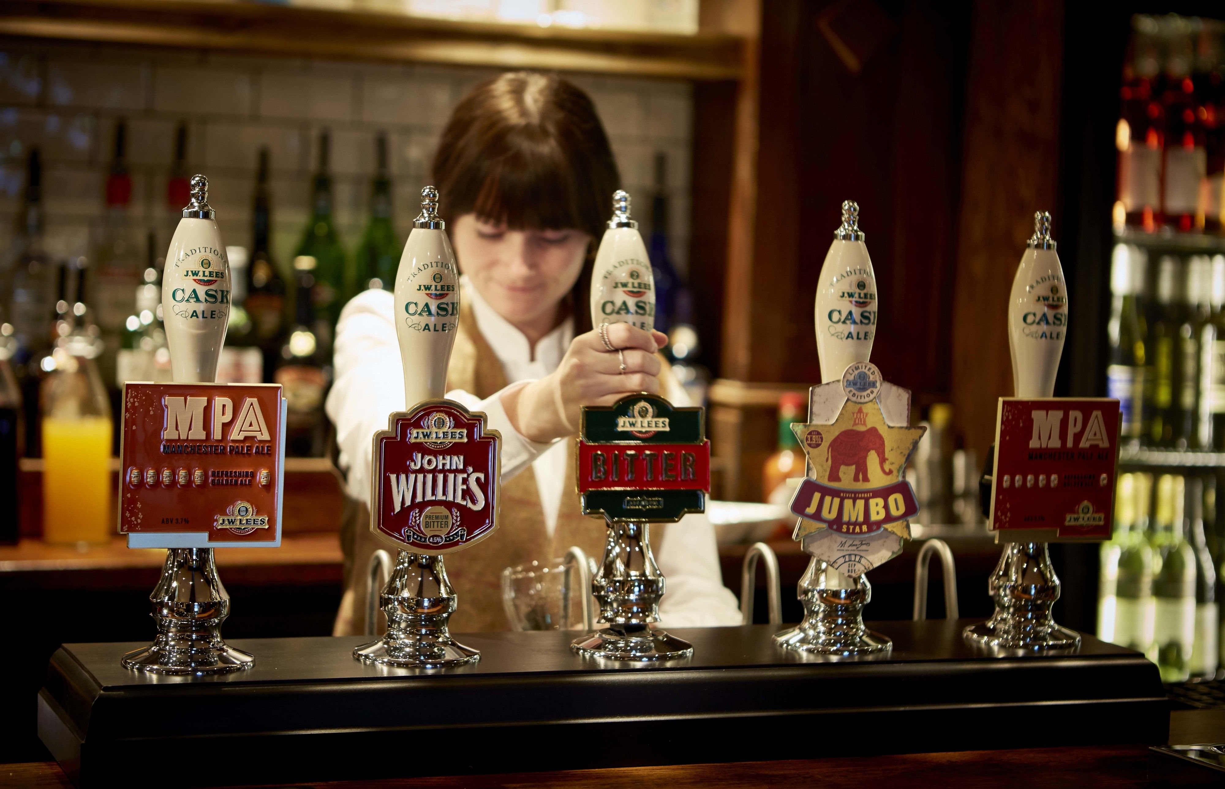 Un bar con múltiples grifos de cerveza, incluyendo marcas como JW Lees Cask Ale y John Willies Bitter.