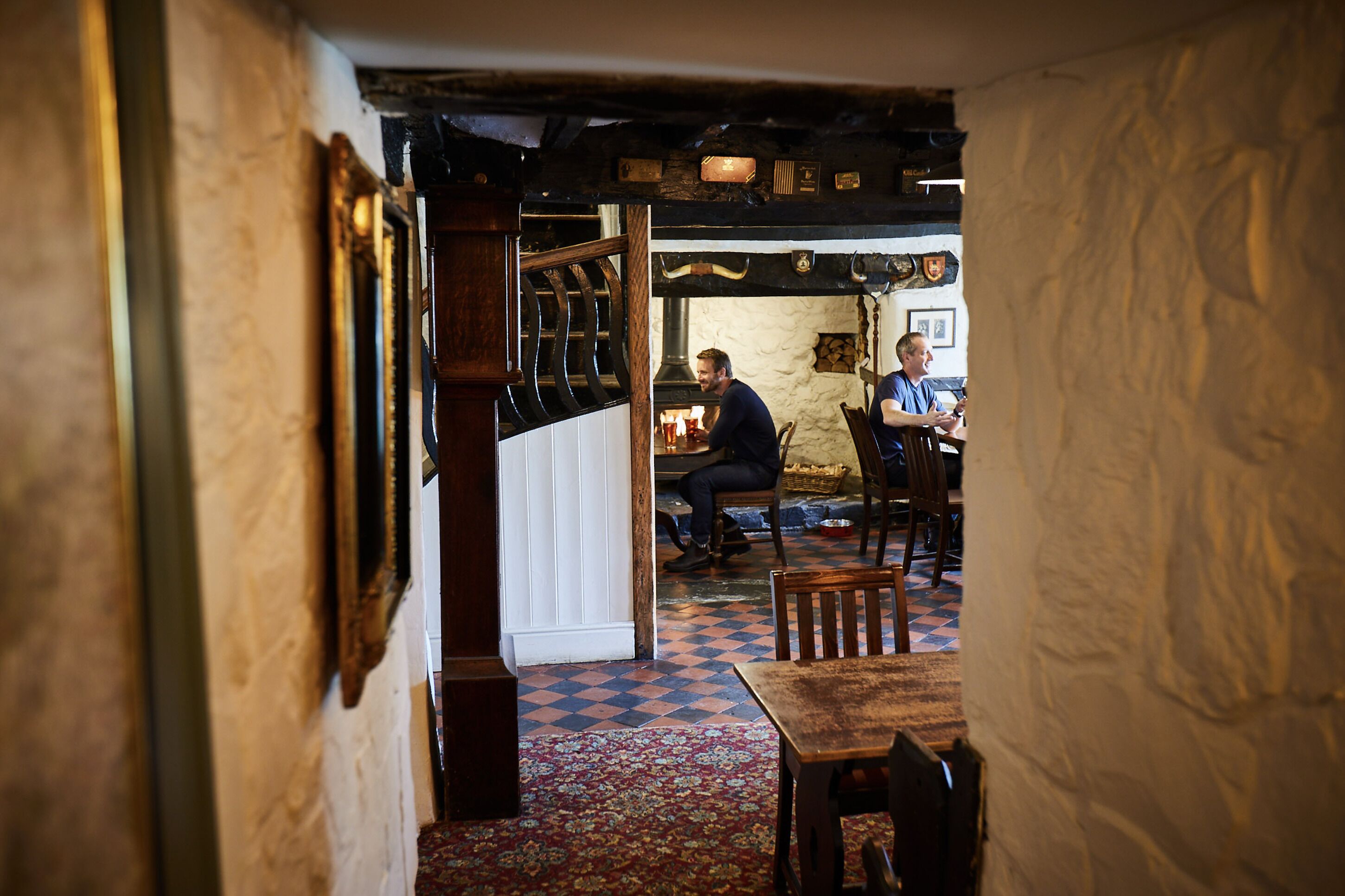 A narrow hallway with a checkered floor leading to a room with a fireplace, wooden furniture, and a stone wall.
