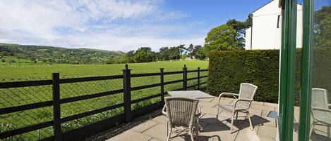 A patio with wicker chairs and a table, overlooking a green field and a white building.