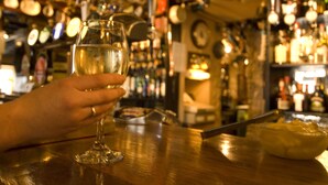 A bar setting with a hand holding a glass of white wine, a knife on the counter, and various bottles in the background. - The Groes Inn (Conwy)