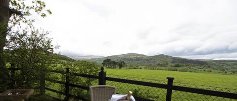 A patio with a table set for two, a stone fountain, and a view of a green field and hills.