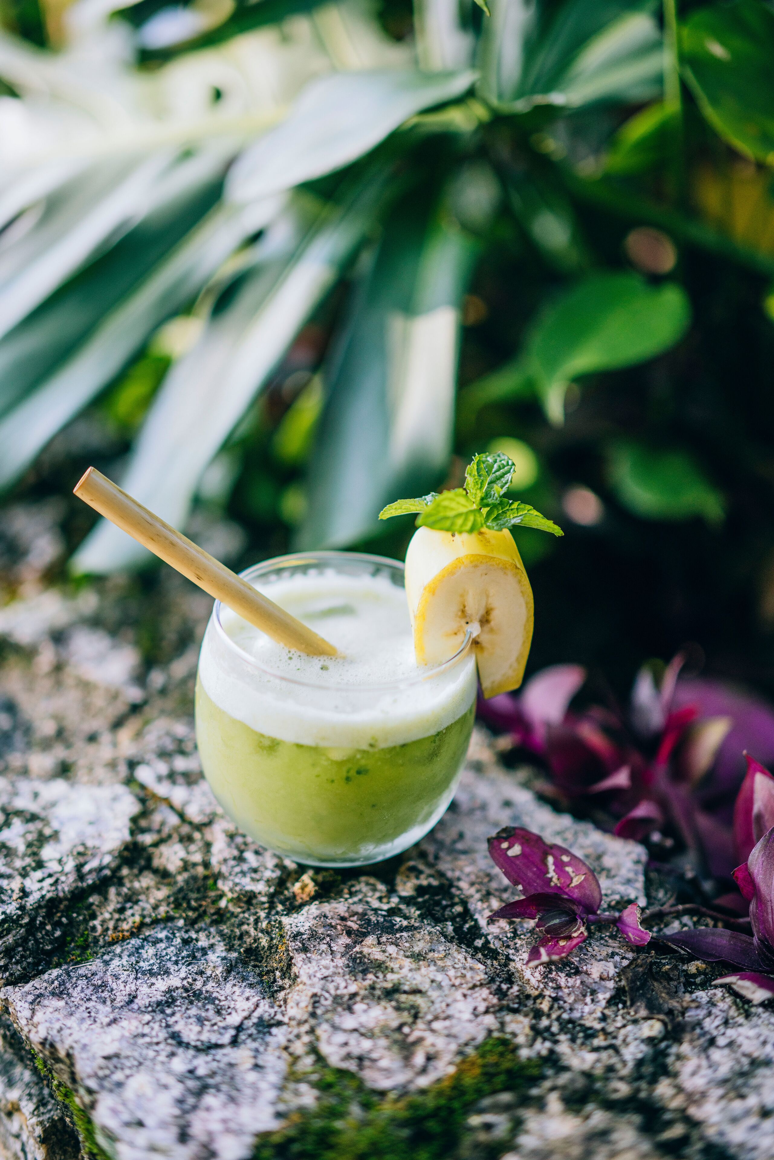 A glass of green beverage with a banana slice and mint garnish on a textured stone surface.