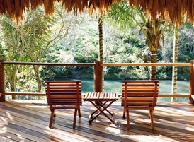 Terraza de madera con dos sillas y una pequeña mesa, con vista a un cuerpo de agua y abundante vegetación.