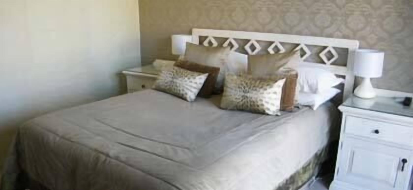 A neatly made bed with a beige bedspread and decorative pillows, a white nightstand with a lamp, and a patterned wallpaper on the wall.