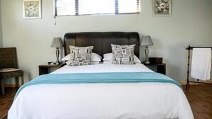 A neatly made bed with patterned pillows, a bedside table with a lamp, and a chair in a room with wooden flooring and a window with blinds.