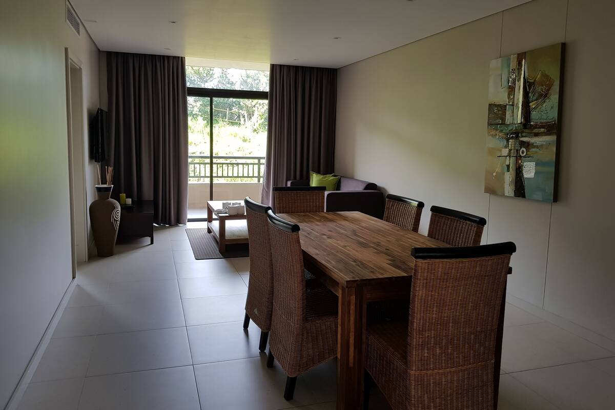 A dining area with a wooden table and wicker chairs, a sofa, a TV, and a painting on the wall.