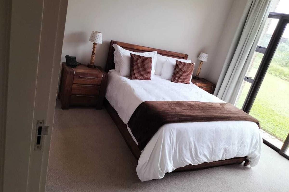 A hotel room with a large bed, wooden headboard, two bedside tables with lamps, and a view of the outdoors through a large window.