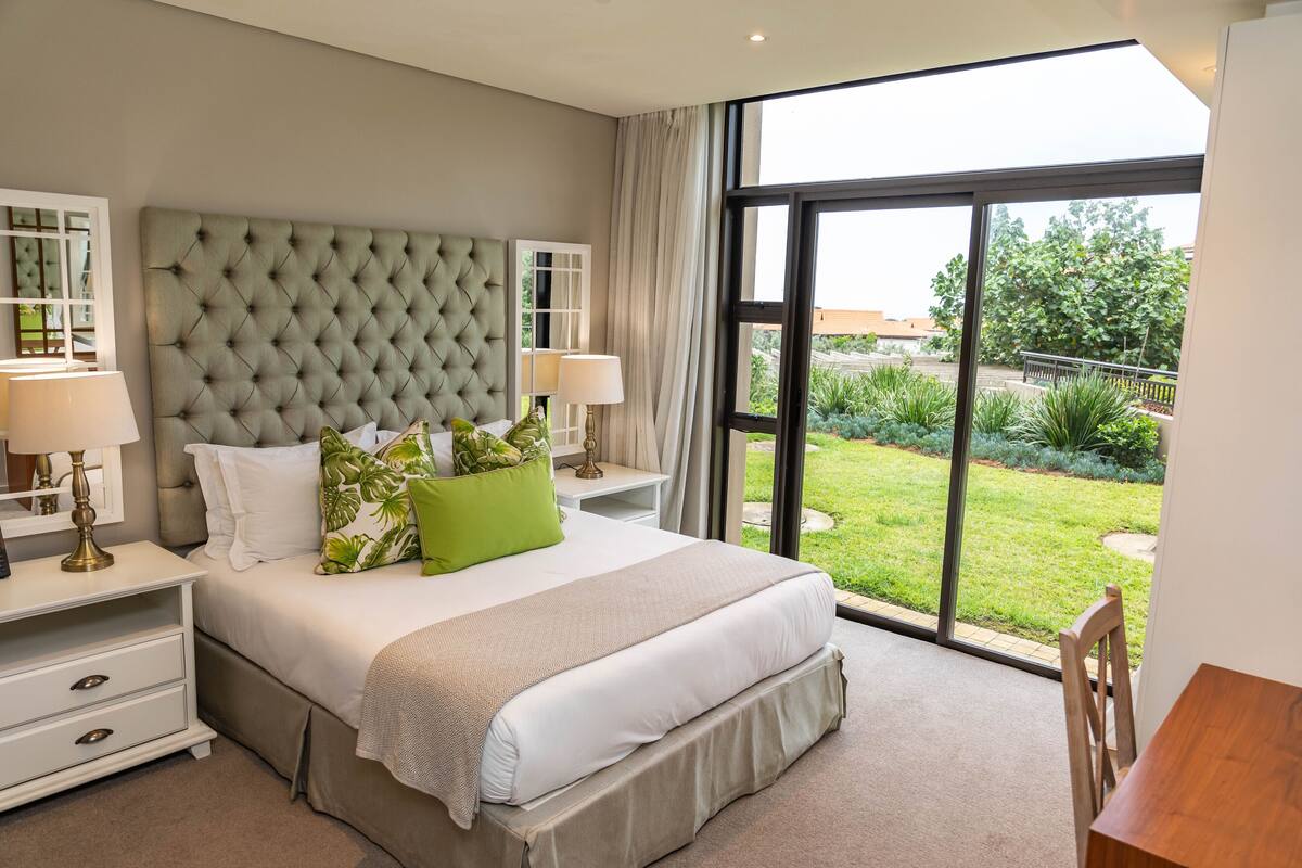 A modern bedroom with a tufted headboard, a large bed with white bedding and green pillows, two bedside tables with lamps, and a view of a garden through a sliding glass door.