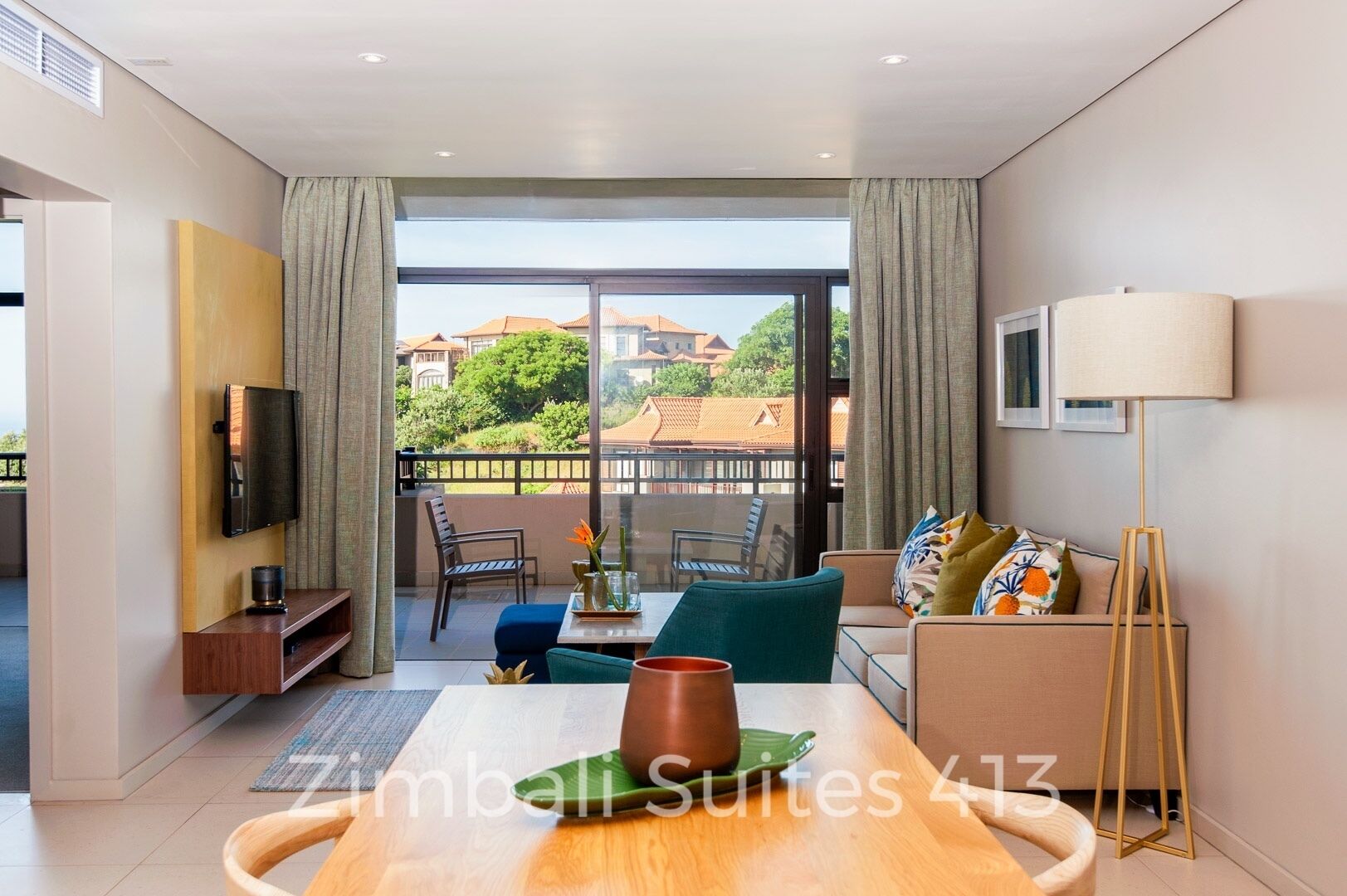 Ein modernes Wohnzimmer mit Balkon, einem flachbildfernseher an der Wand, einem Esstisch mit Stühlen und einer Couch mit Kissen.