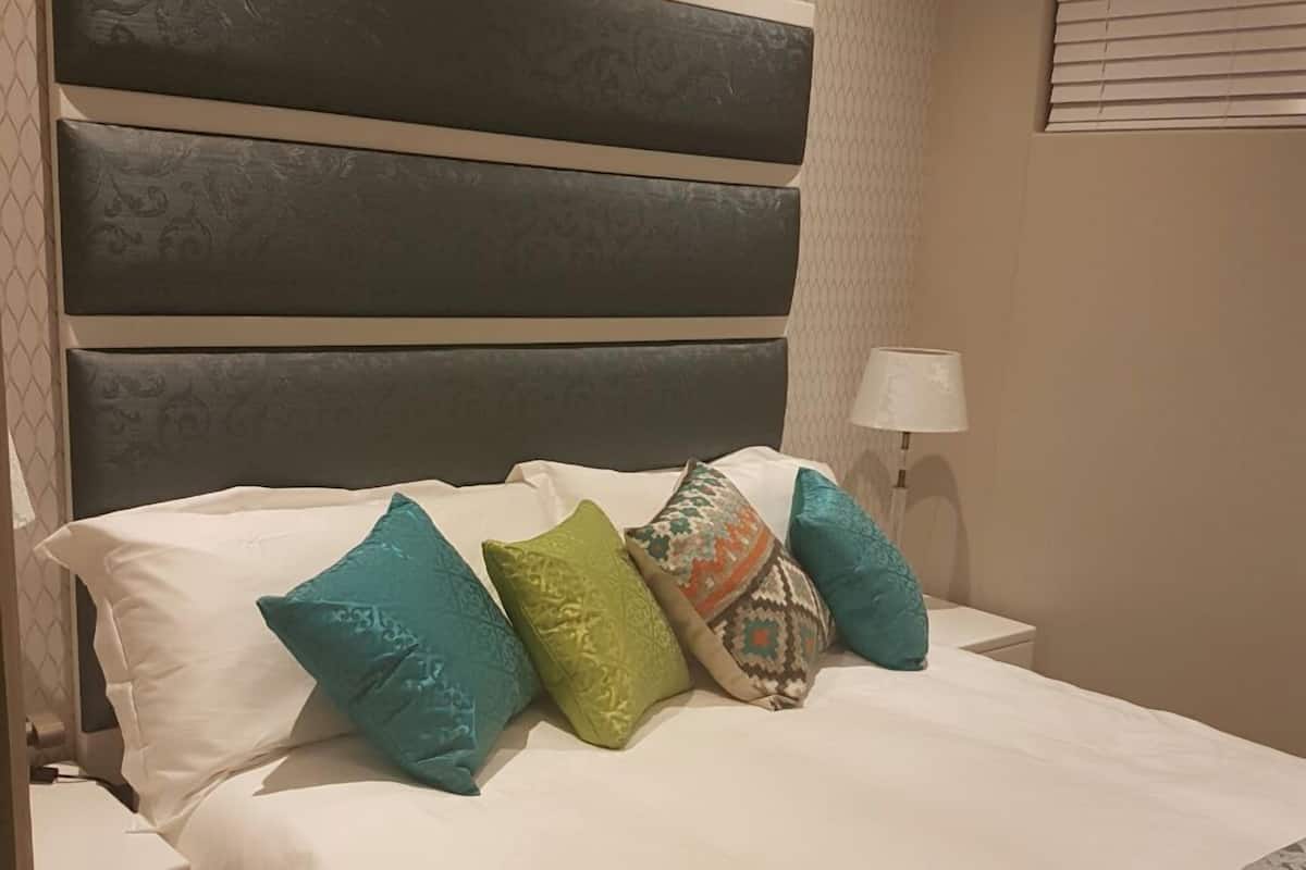 A neatly made bed with white bedding and multiple colorful pillows, a headboard with a textured pattern, and a lamp on the nightstand.