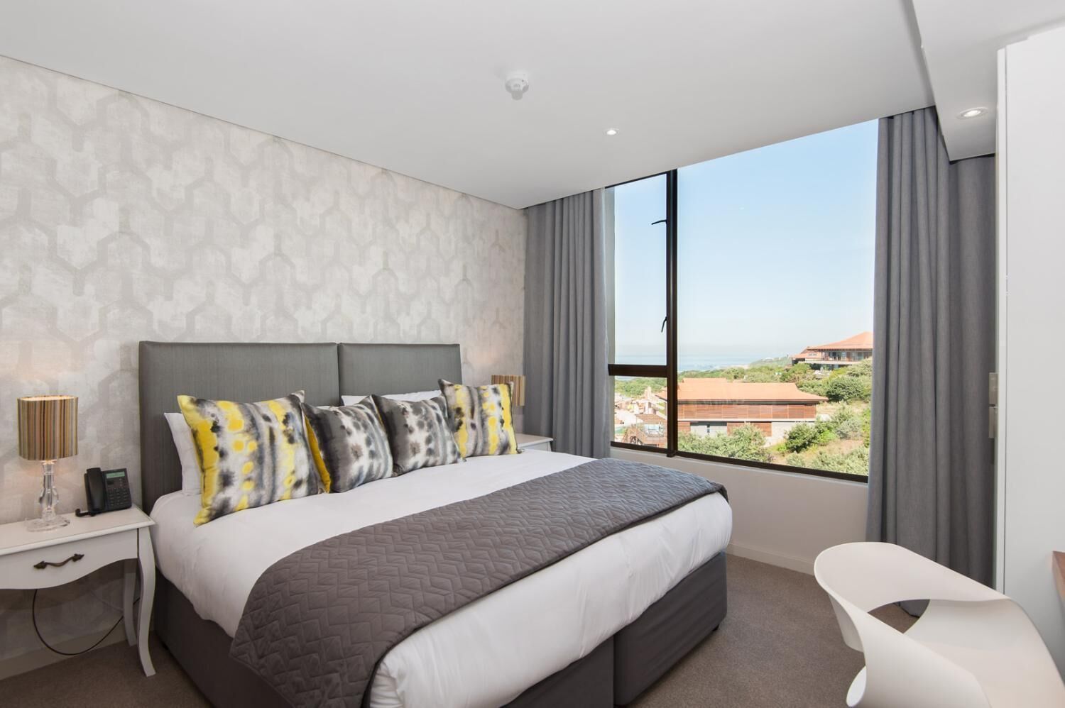 Una habitación de hotel moderna con una cama grande, una mesita de noche con lámpara, una silla y vistas a la ciudad a través de amplias ventanas.