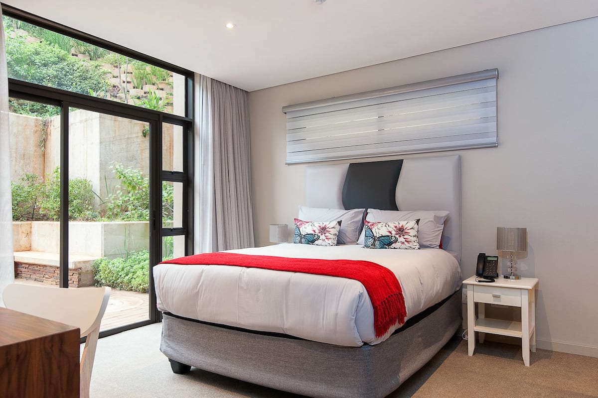 A modern bedroom with a large bed, a bedside table with a lamp, and a view of greenery through large windows.