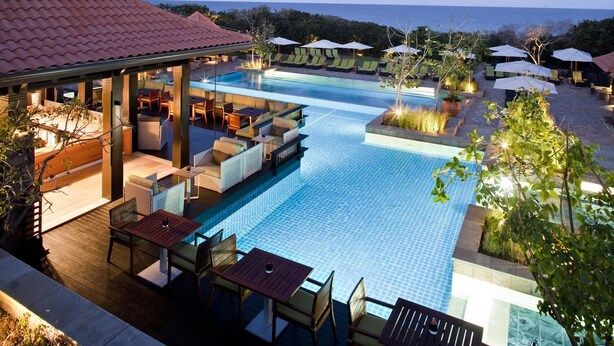 Área de piscina no terraço com espreguiçadeiras e guarda-sóis, rodeada por um deck de madeira e vista para o oceano.