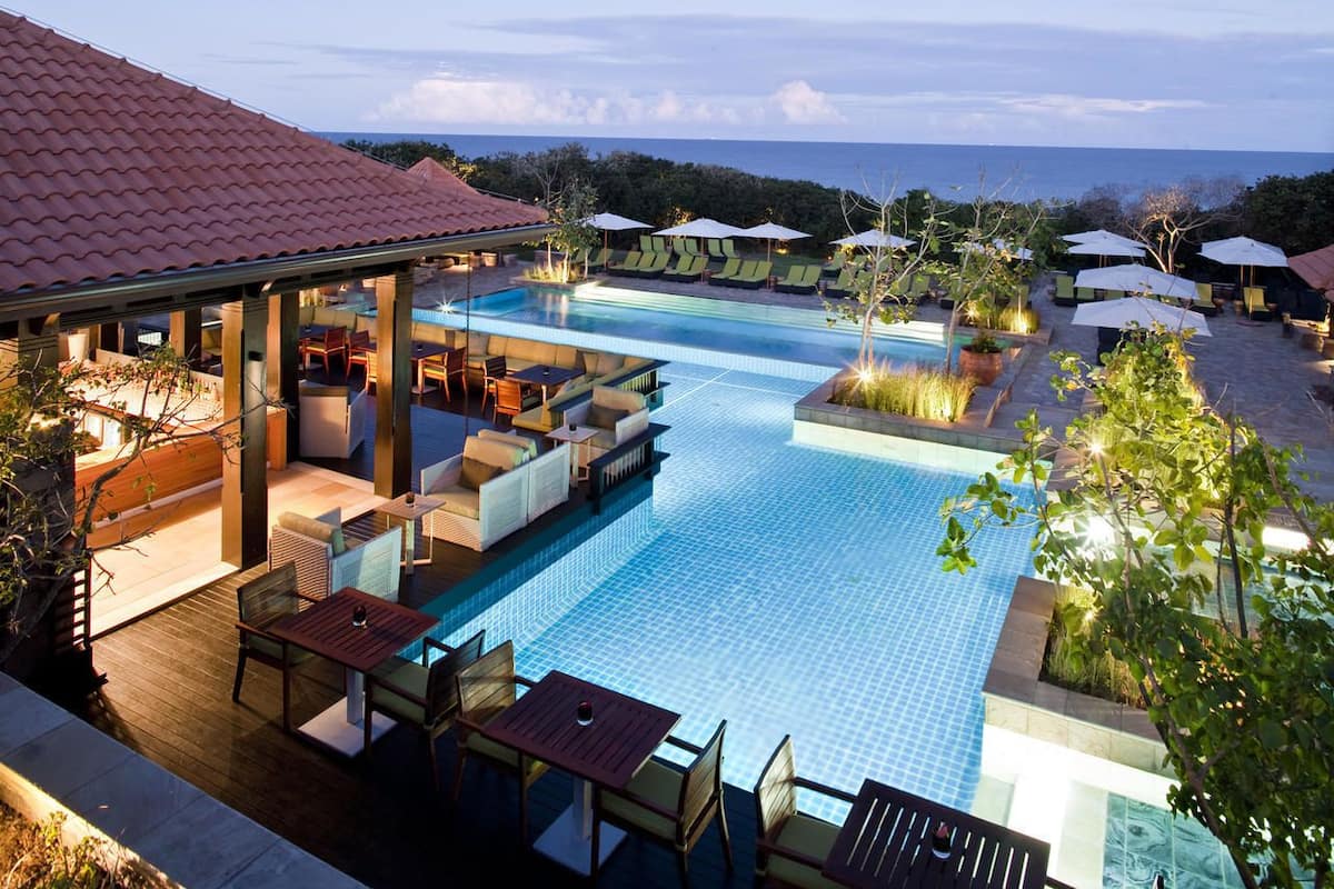 A rooftop pool area with lounge chairs and umbrellas, surrounded by a wooden deck and a view of the ocean.