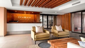 Um interior moderno com uma mesa de recepção identificada como "ZIMBAI SUITES", dois sofás, uma mesa de centro e uma janela ampla com vista.