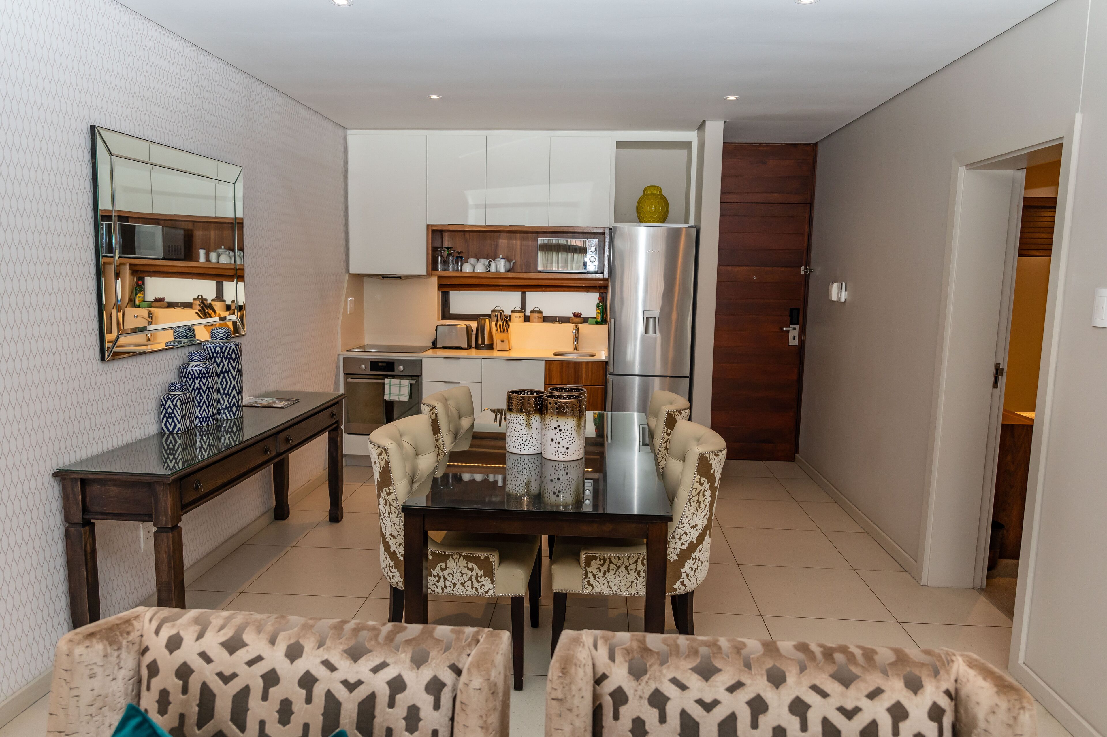 A modern kitchen with white cabinets, a stainless steel refrigerator, and a dining area with patterned chairs and a glass table.