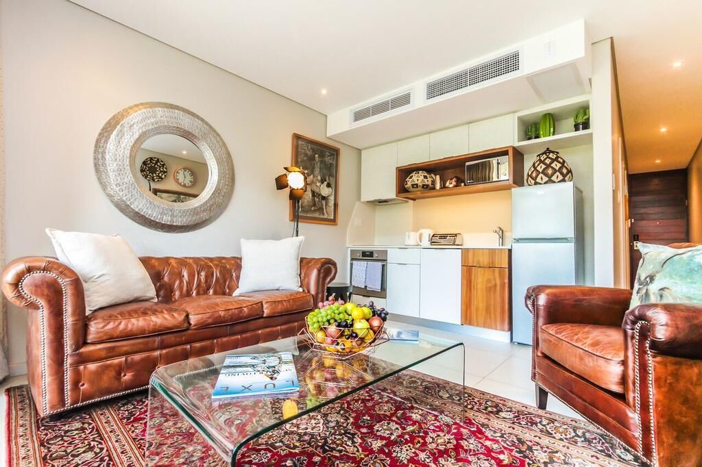 Un salón con un sofá de cuero marrón, una mesa de centro de vidrio y un tapete rojo estampado. Hay una zona de cocina con armarios blancos y un refrigerador.
