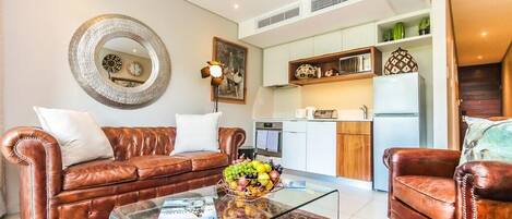 Un salón con un sofá de cuero marrón, una mesa de centro de vidrio y un tapete rojo estampado. Hay una zona de cocina con armarios blancos y un refrigerador.