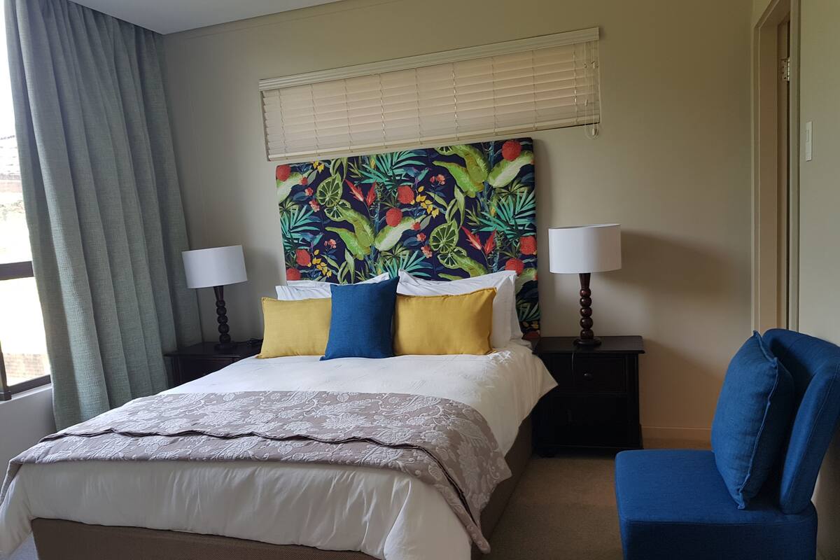 A hotel room with a bed, two bedside tables, a chair, a window with curtains, and a colorful tropical-themed headboard.