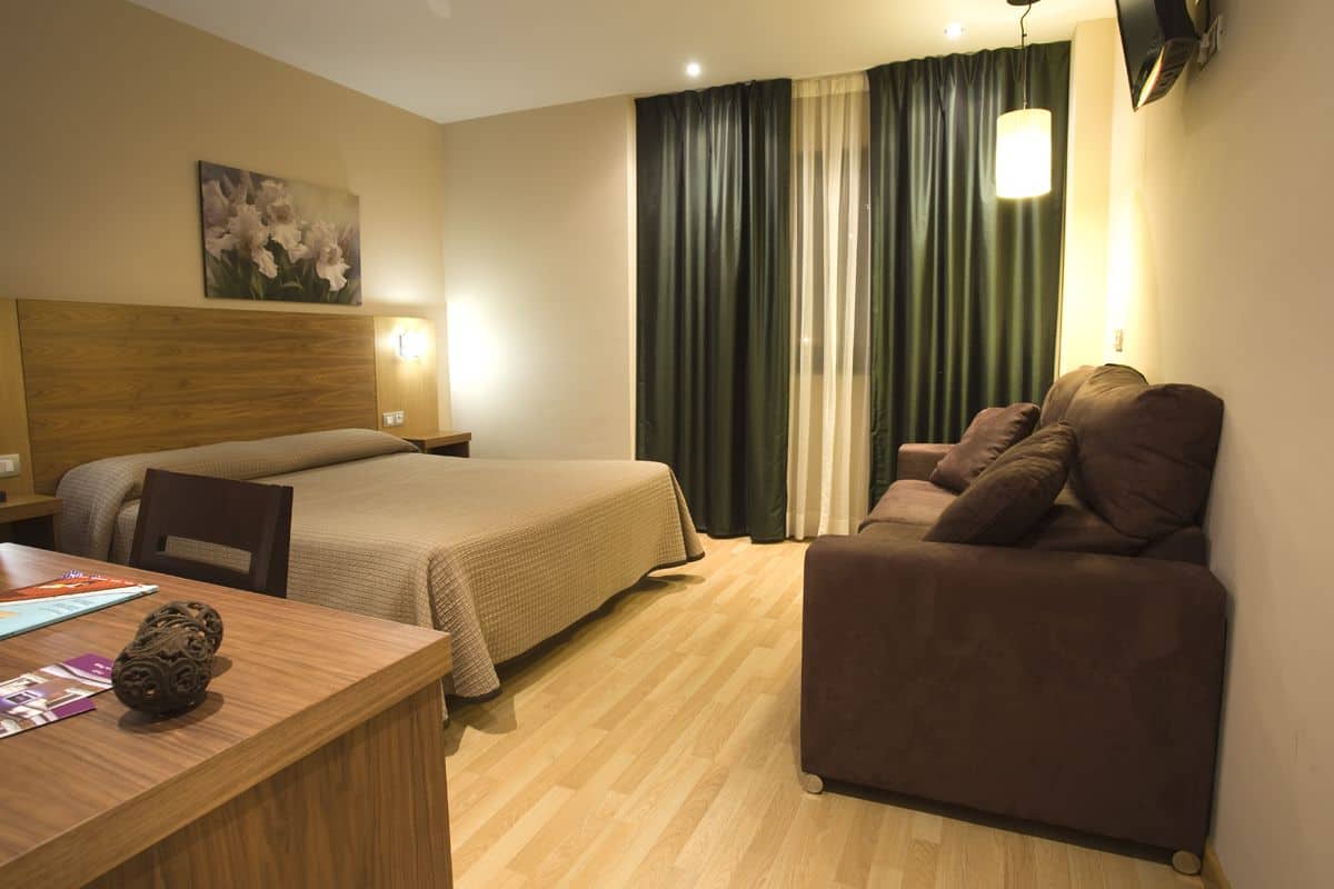 Quarto de hotel com cama de cabeceira de madeira, um sofá, uma escrivaninha com abajur e um quadro na parede.