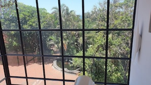 A room with a large window featuring a view of a garden with various trees and a small pond. - Zimbali beach estate  (Ballito)