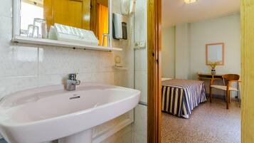 Ein Badezimmer mit Waschbecken, Fön und Spiegel. Im Hintergrund ist ein Bett mit gestreifter Bettdecke zu sehen.
