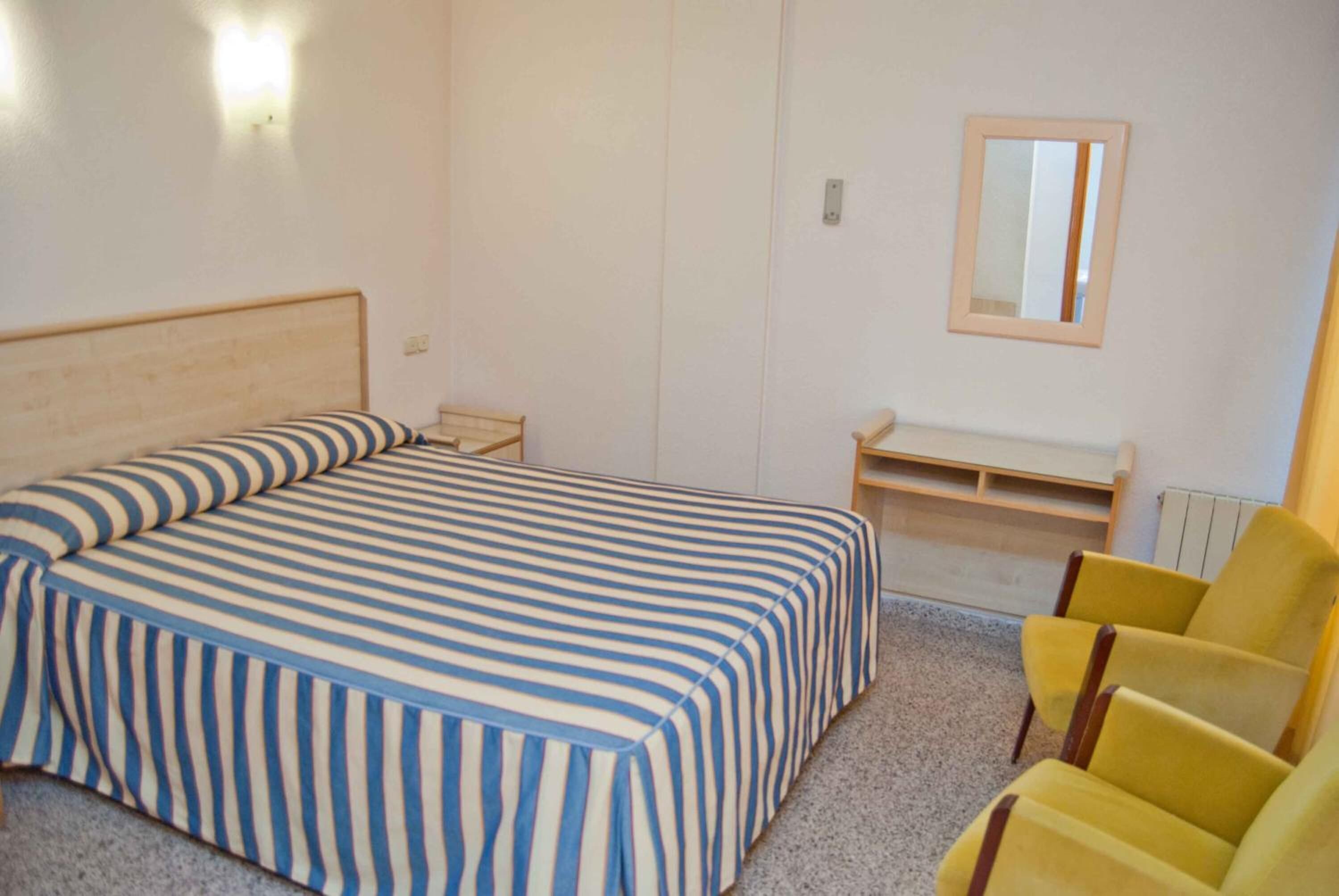 A bedroom with a bed, a bedside table, a chair, a desk, and a mirror.