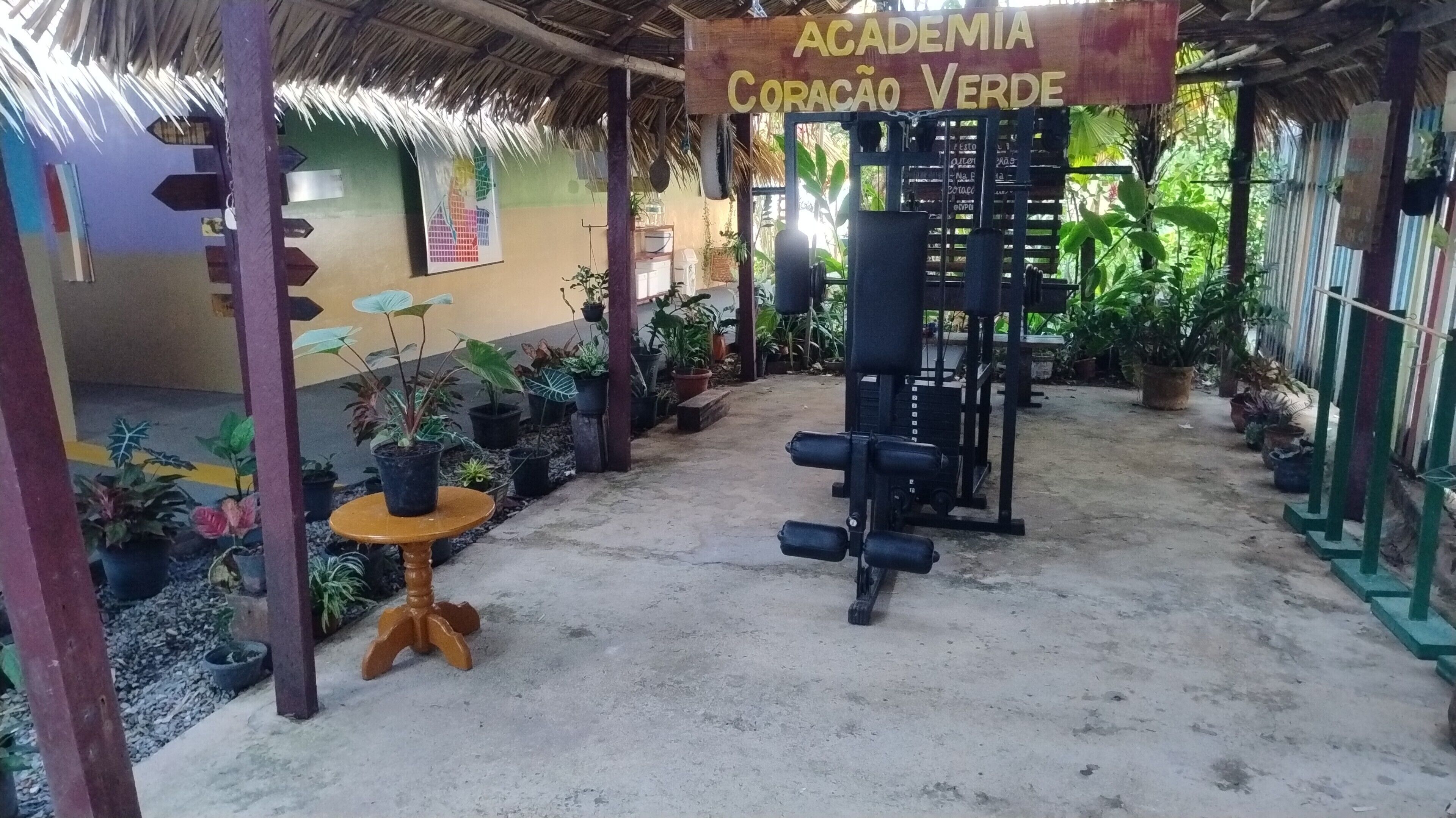 An outdoor gym area with exercise equipment, surrounded by potted plants and a sign reading 'Academia Coracao Verde'.