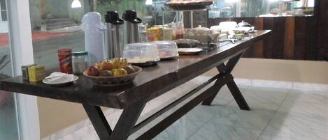 Una mesa de buffet con diferentes alimentos y salsas, ubicada en un espacio con ventanas amplias y piso de baldosas.
