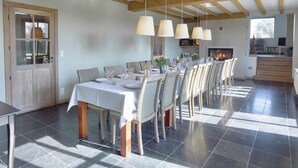 Een eethoek met een lange tafel gedekt voor zes personen, houten balken in het plafond en een open haard op de achtergrond.