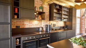 Een keuken met donkere kastjes, een bakstenen muur en een houten plafond. Er staan planken met glazen en er staat een koffieapparaat.