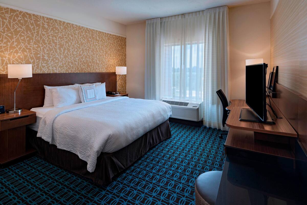 A hotel room with a large bed, a desk with a flat-screen TV, a desk lamp, and a window with curtains.