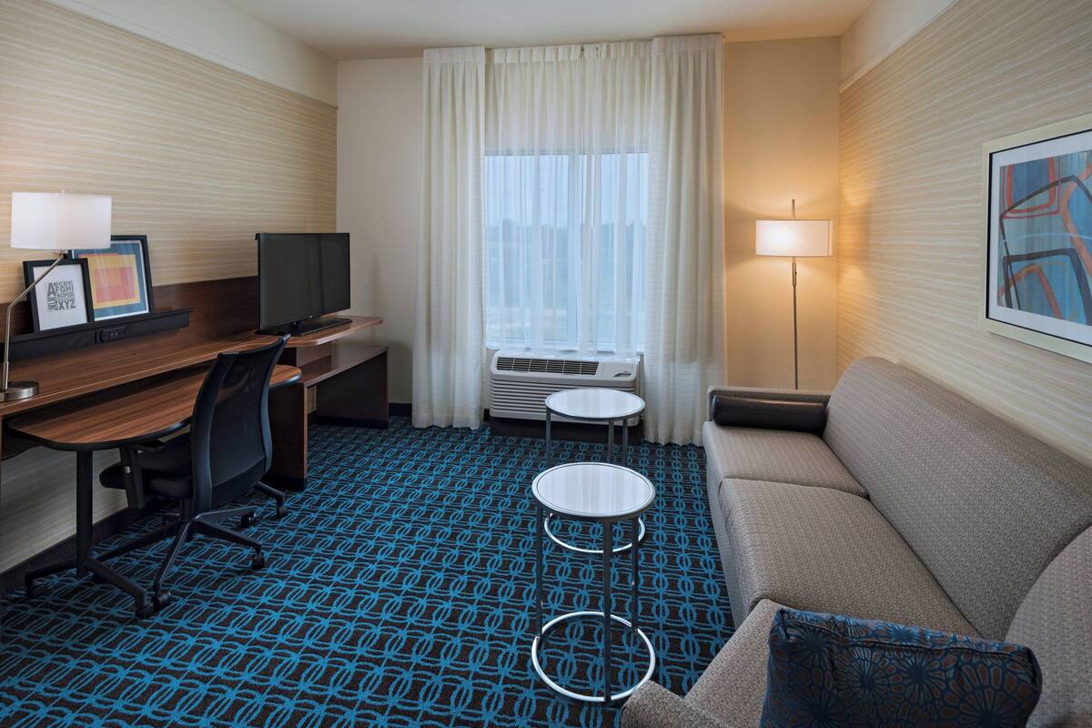 A hotel room with a desk, chair, sofa, two small tables, a TV, and a large window with curtains.
