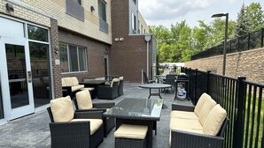 Une terrasse extérieure aménagée avec des meubles en osier et une table en verre, entourée d’immeubles en briques et d’un mur de pierre.