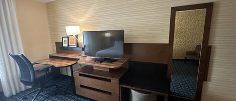 A hotel room with a wooden desk, a television, a chair, a lamp, and a mirror.