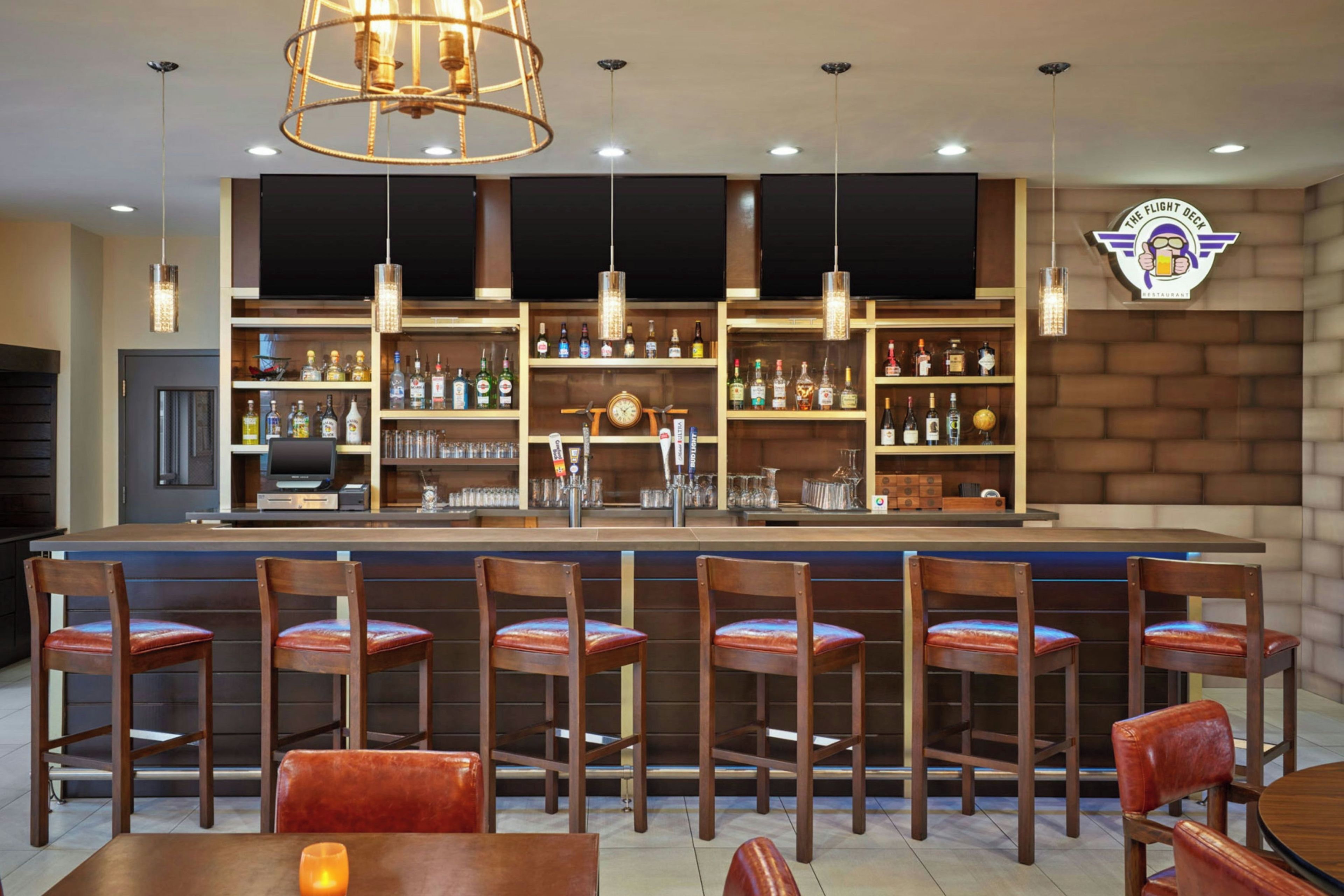 A bar area with multiple seating options, a counter with a cash register, and a variety of bottles and glasses on shelves.