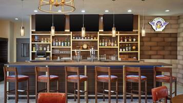 A bar area with multiple seating options, a counter with a cash register, and a variety of bottles and glasses on shelves.