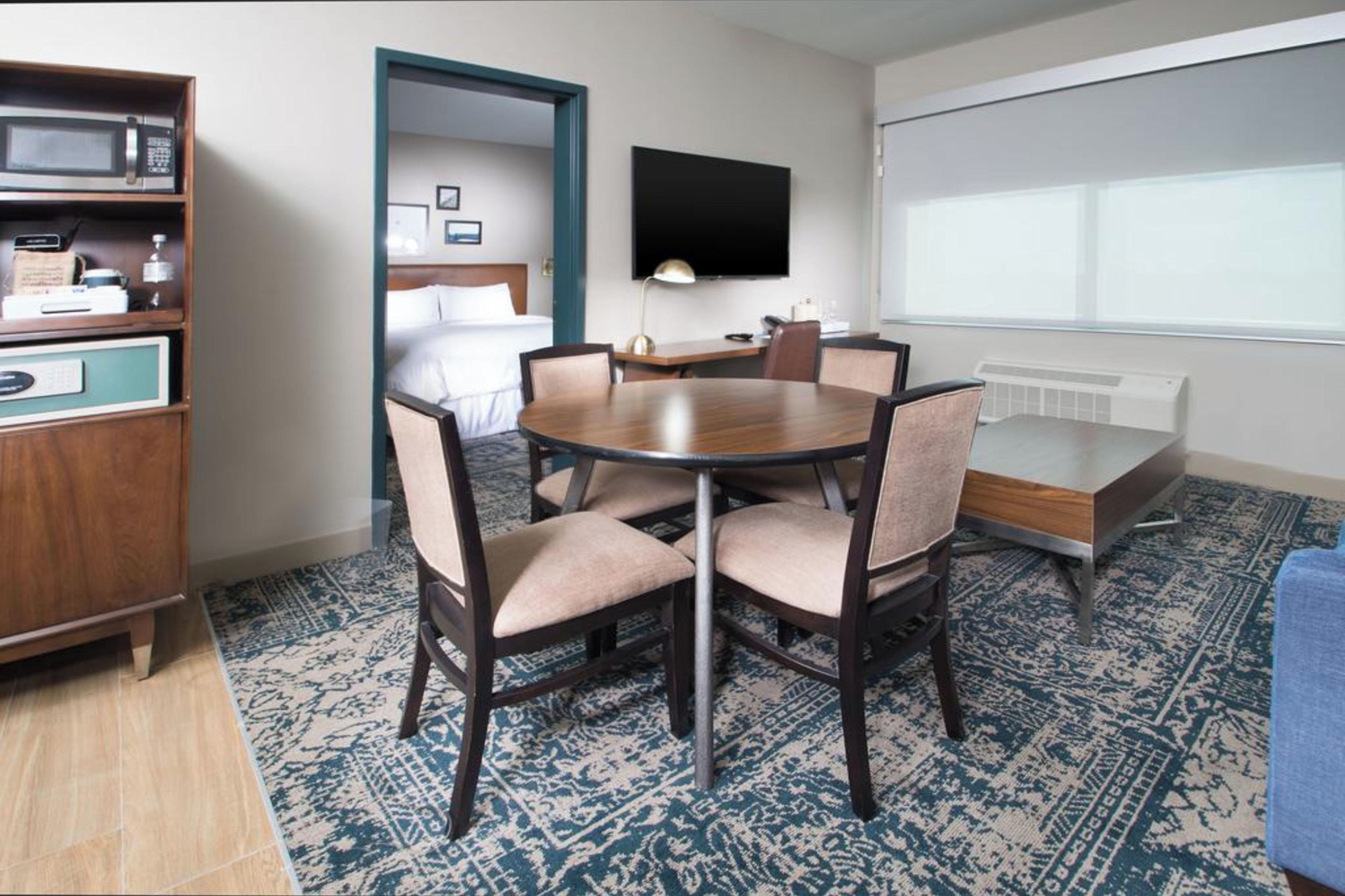 Una habitación de hotel con una mesa redonda, cuatro sillas, una cama, un televisor y un minibar.