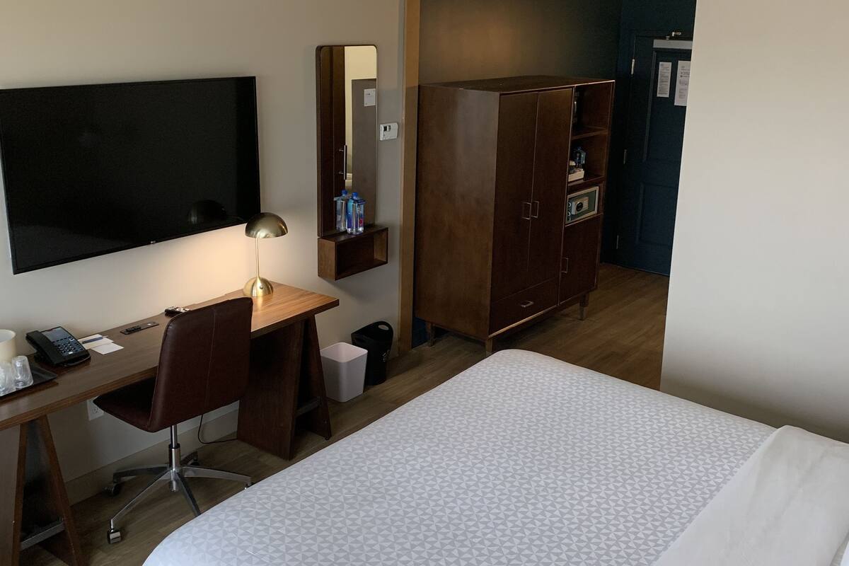 Een hotelkamer met een bed, bureau, stoel, televisie, kast en een kleine prullenbak.
