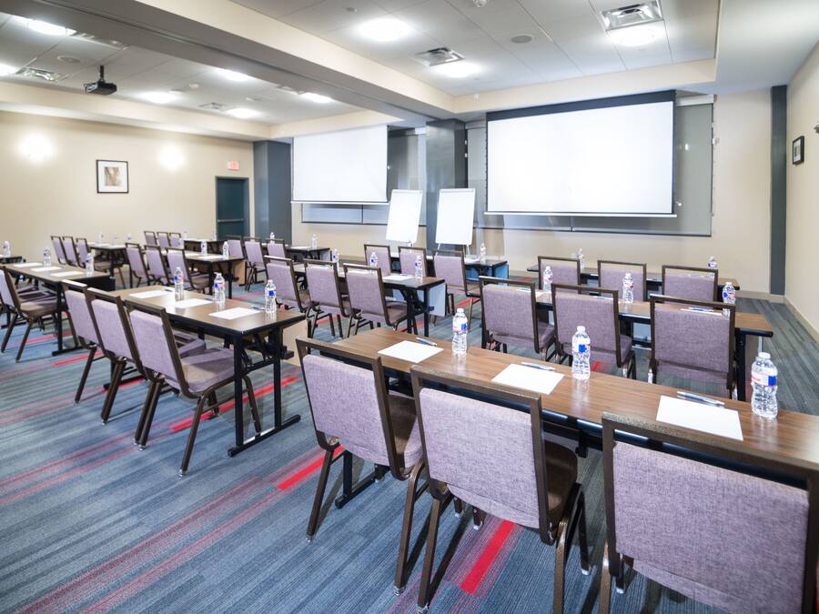 Uma sala de conferências com fileiras de mesas e cadeiras, equipada com garrafas de água e uma tela grande para apresentações.